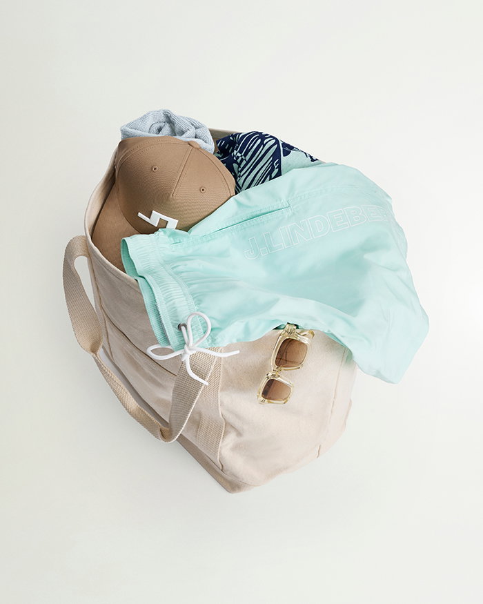 flat lay image of a tote bag filled with JL apparel including swim shorts a cap sunglasses and other summer knits