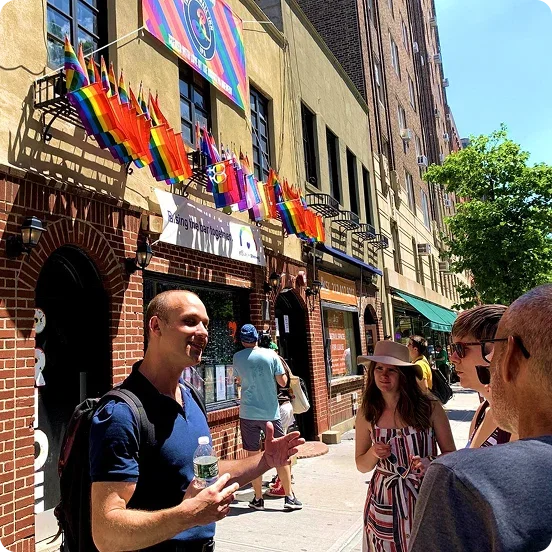 A tour guide speaking to a small group on a city sidewalk in front of a brick building decorated with rainbow flags.