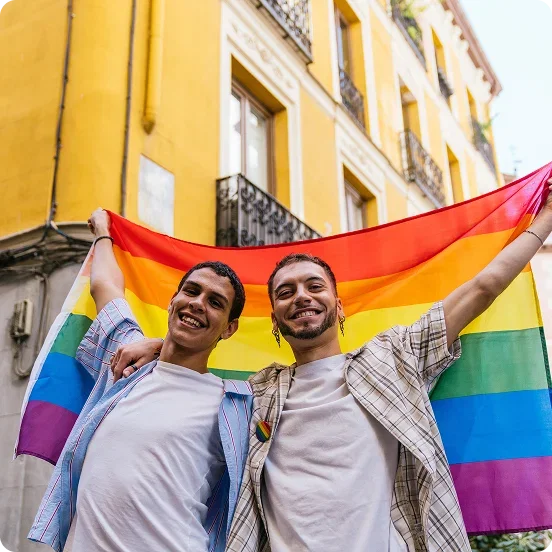 Two smiling young men standing together on a city street and holding up a large rainbow Pride flag.