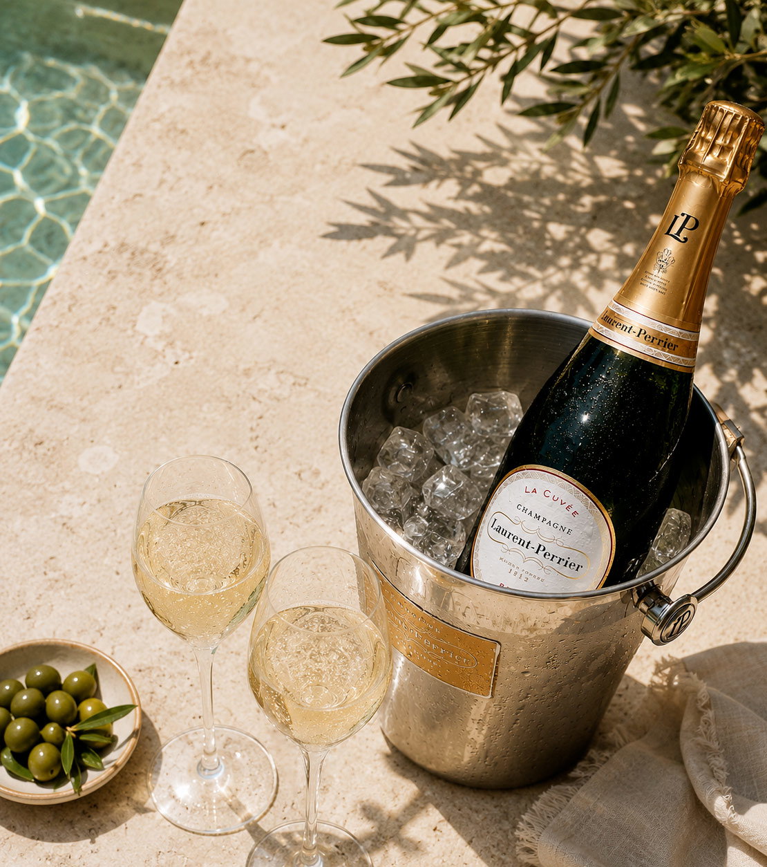 Poolside Laurent-Perrier champagne bottle chilling in an ice bucket with two filled flutes and a bowl of green olives on a stone deck