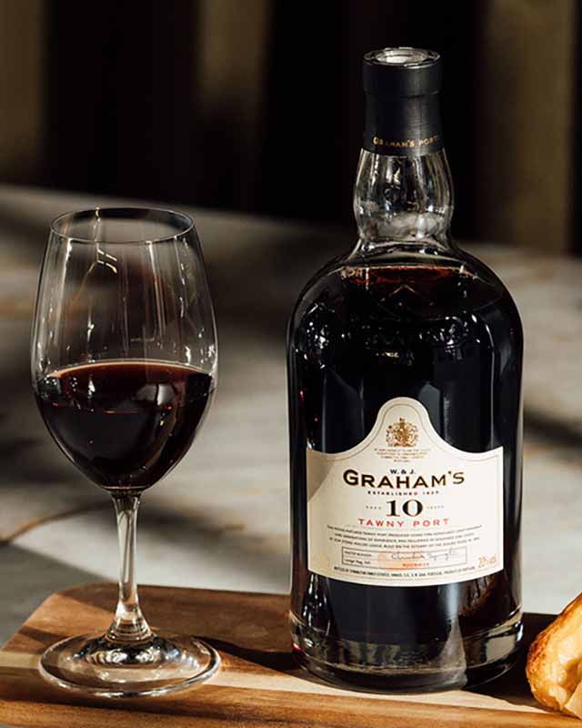 A bottle of Graham's 10 Tawny Port wine next to a filled wine glass on a wooden surface.