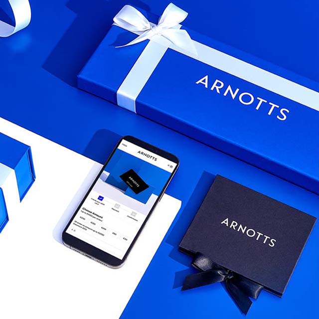 A phone displaying the Arnotts e gift card and a physical gift card in a gift box, surrounded by blue gift boxes with white and blue ribbons.