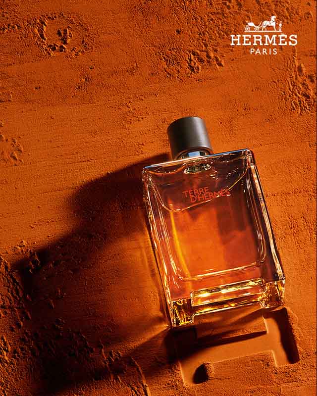 A bottle of Terre d'Hermès perfume lies on a textured, earthy orange surface with the Hermès logo above.