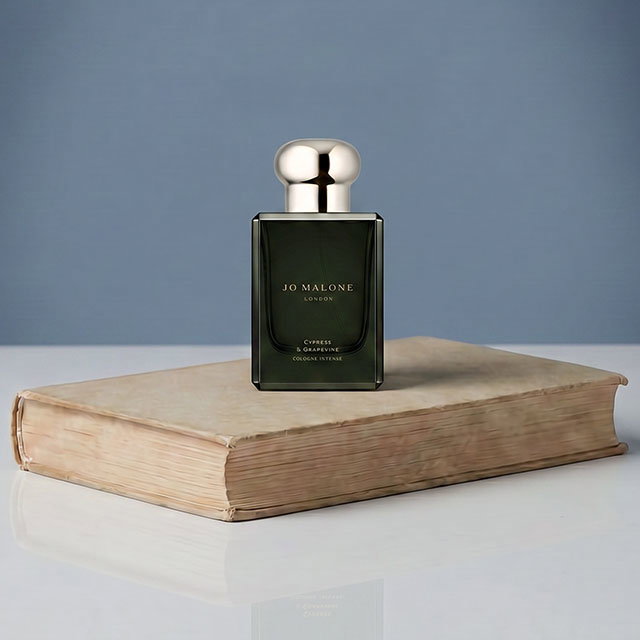 A bottle of Jo Malone perfume rests on an old hardcover book against a plain background.