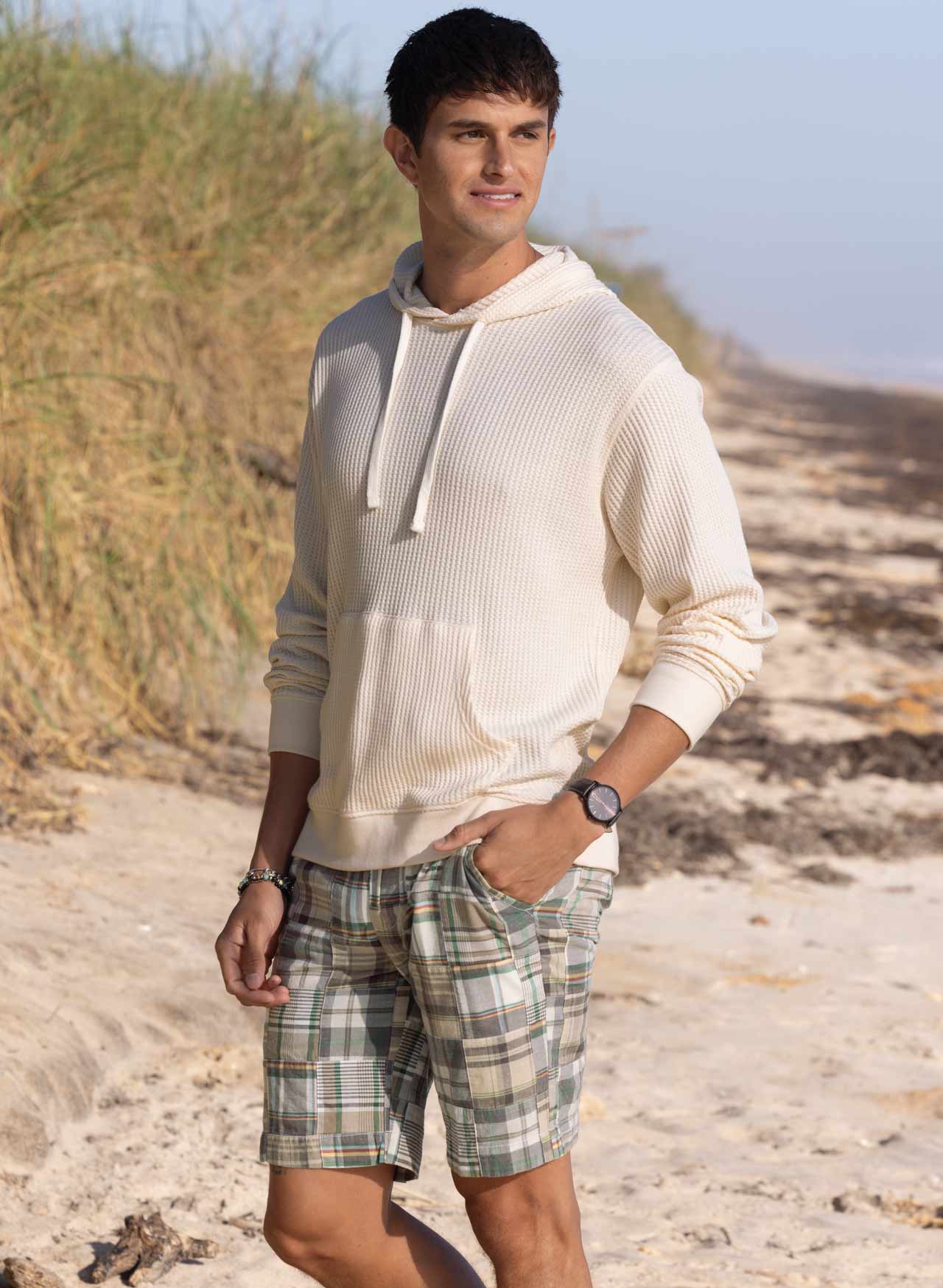 Man walking on beach wearing plaid madras shorts and a cream waffle-knit hoodie