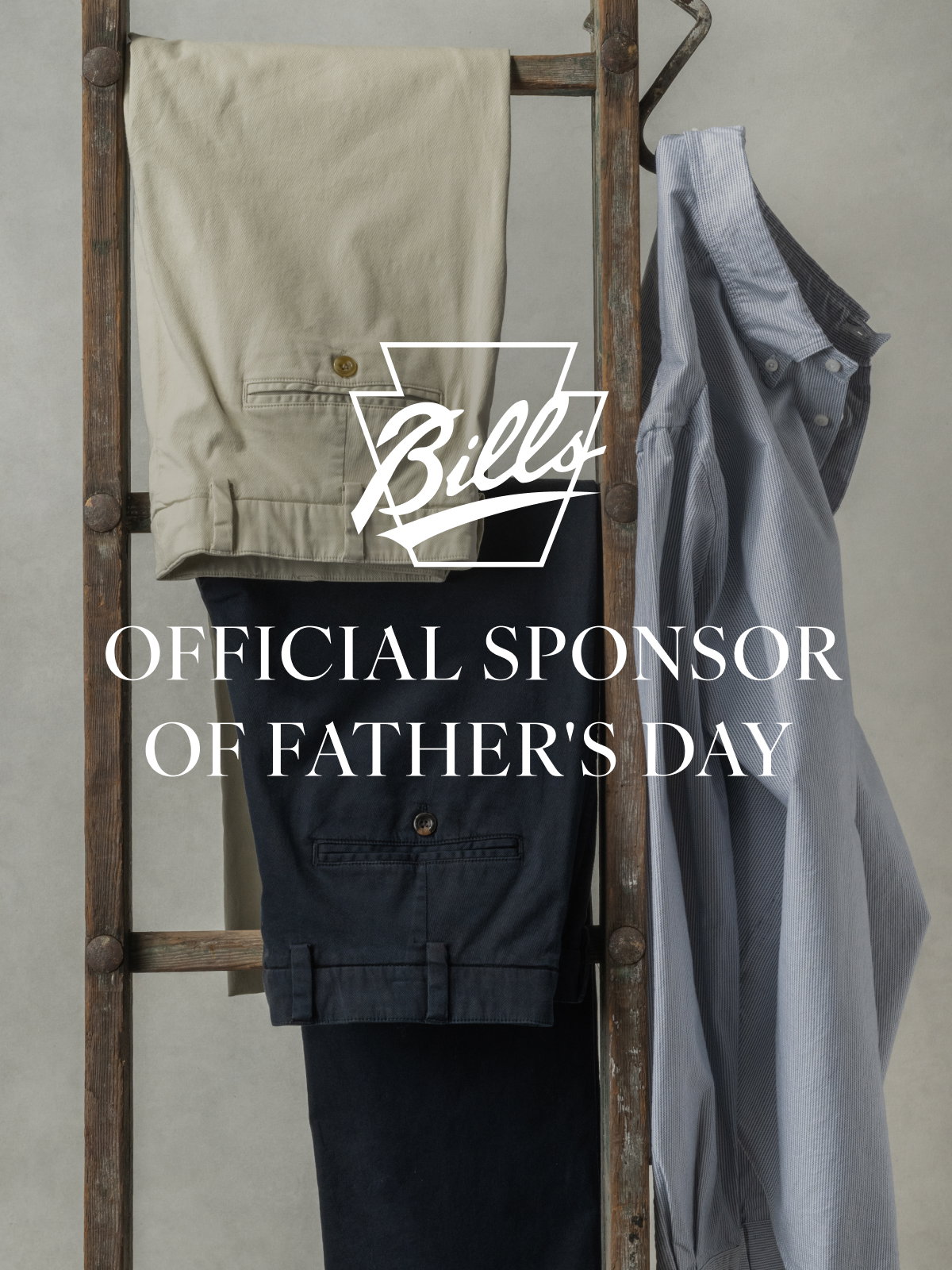 Two pairs of Bills Khakis pants and a Bills Khakis Standard fit shirt hang from a vintage ladder. Text overlay has the Bills Khakis logo and text reads 'Official Sponsor of Father's Day'