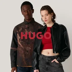 Zwei Models stehen vor einem neutralen Hintergrund. Eines trägt ein gemustertes braunes Hemd und blaue Jeans, das andere eine schwarze cropped Jacke und graue Hosen.