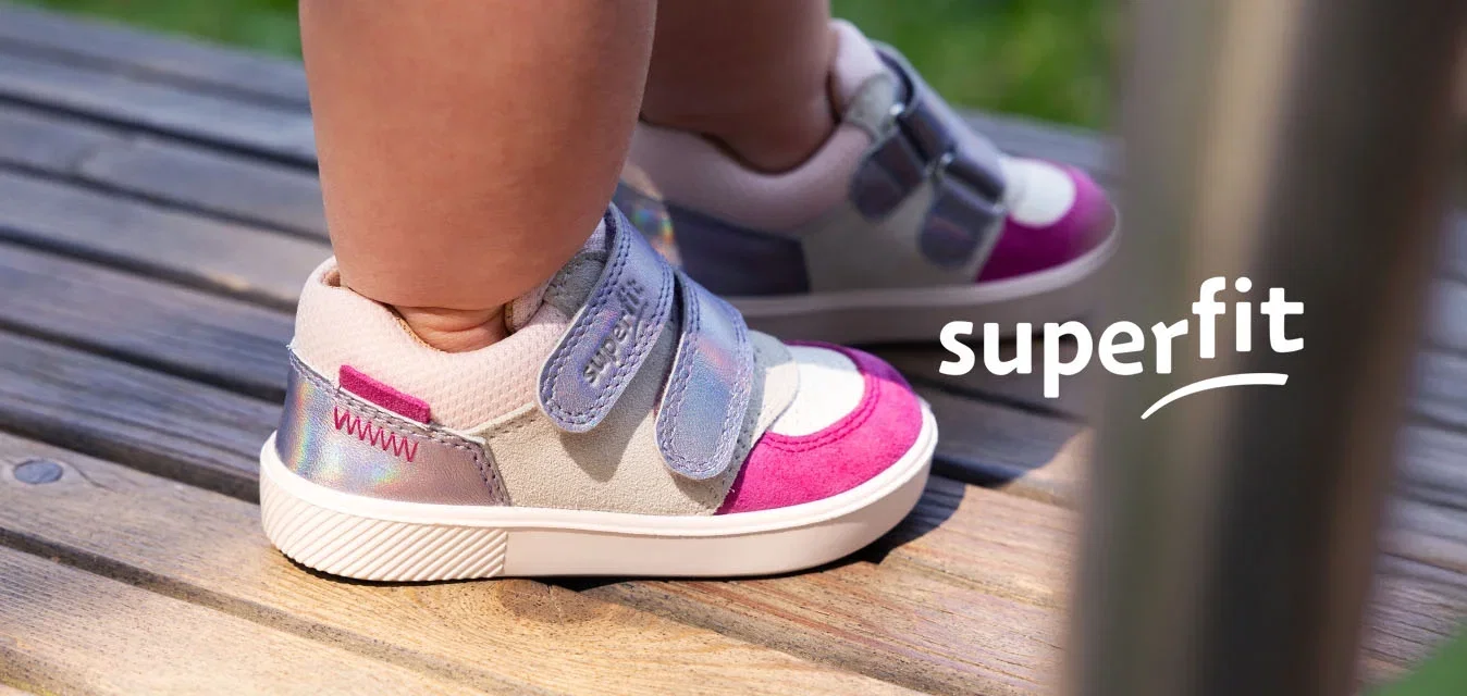Kleinkind trägt lila, pinke und weiße Sneakers und steht auf einem hölzernen Deck im Freien, mit verschwommenem Gras im Hintergrund.