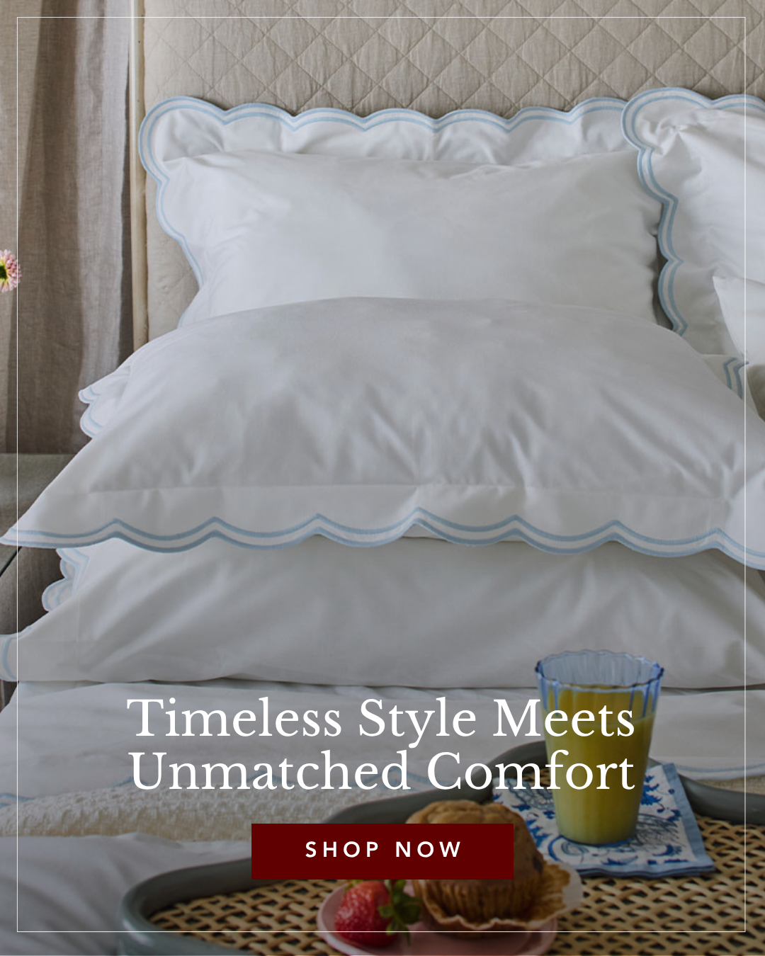 A neatly made bed with white bedding and light blue scalloped trim on pillows and duvet. A quilted headboard is visible behind. A tray with orange juice, a muff