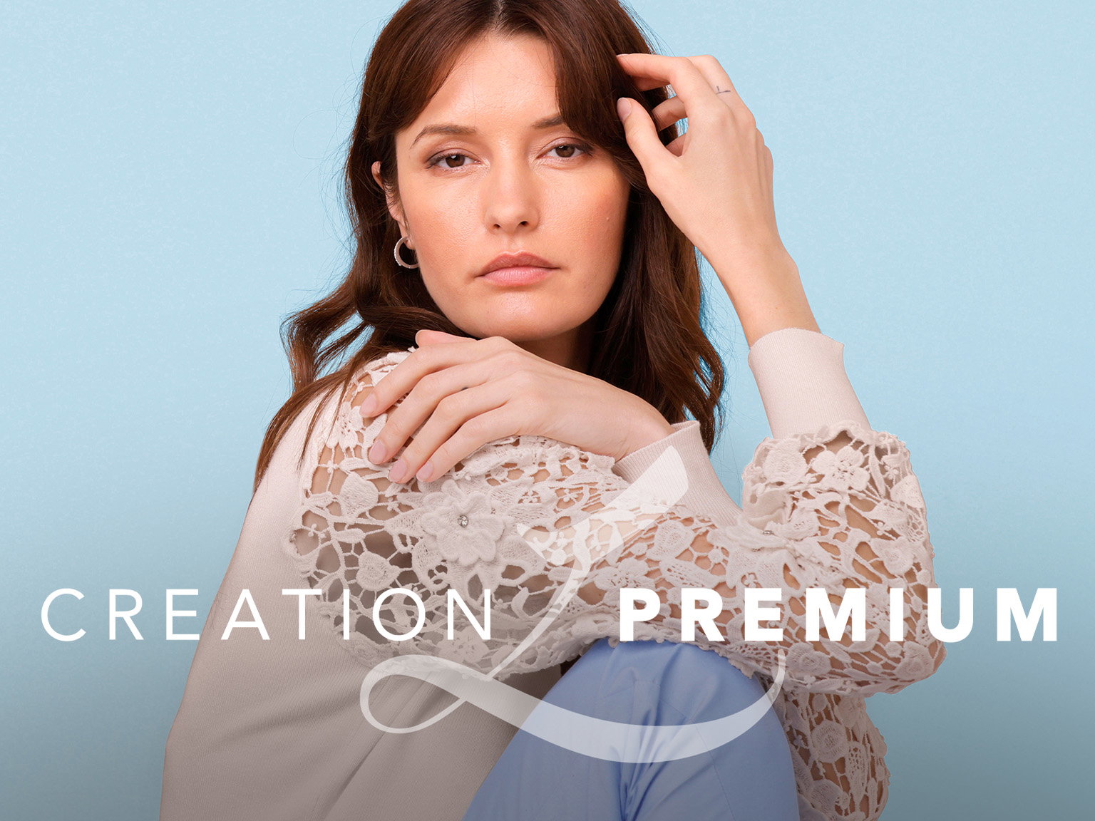 Das Bild zeigt eine Frau in eleganter, cremefarbener Spitzenbluse und blauer Hose, die entspannt vor hellblauem Hintergrund sitzt. Mittig ist das 'CREATION L PREMIUM' Logo in weißer Schrift platziert.