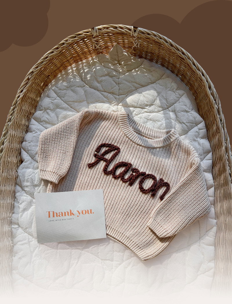 Flat lay of a personalized knit sweater, soft blanket, and thank-you card arranged inside a woven baby basket.