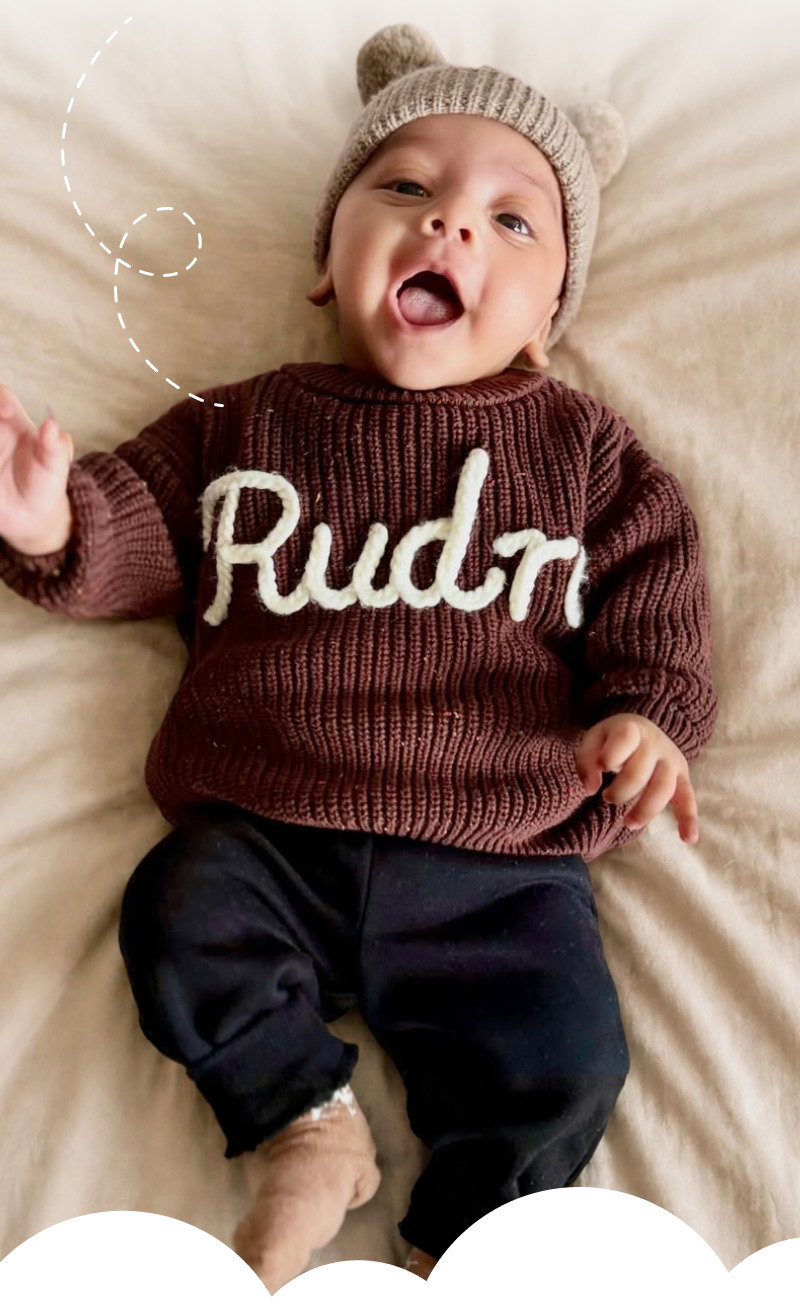 Smiling baby wearing a brown personalized knit sweater and knit hat while lying on a soft cream blanket.