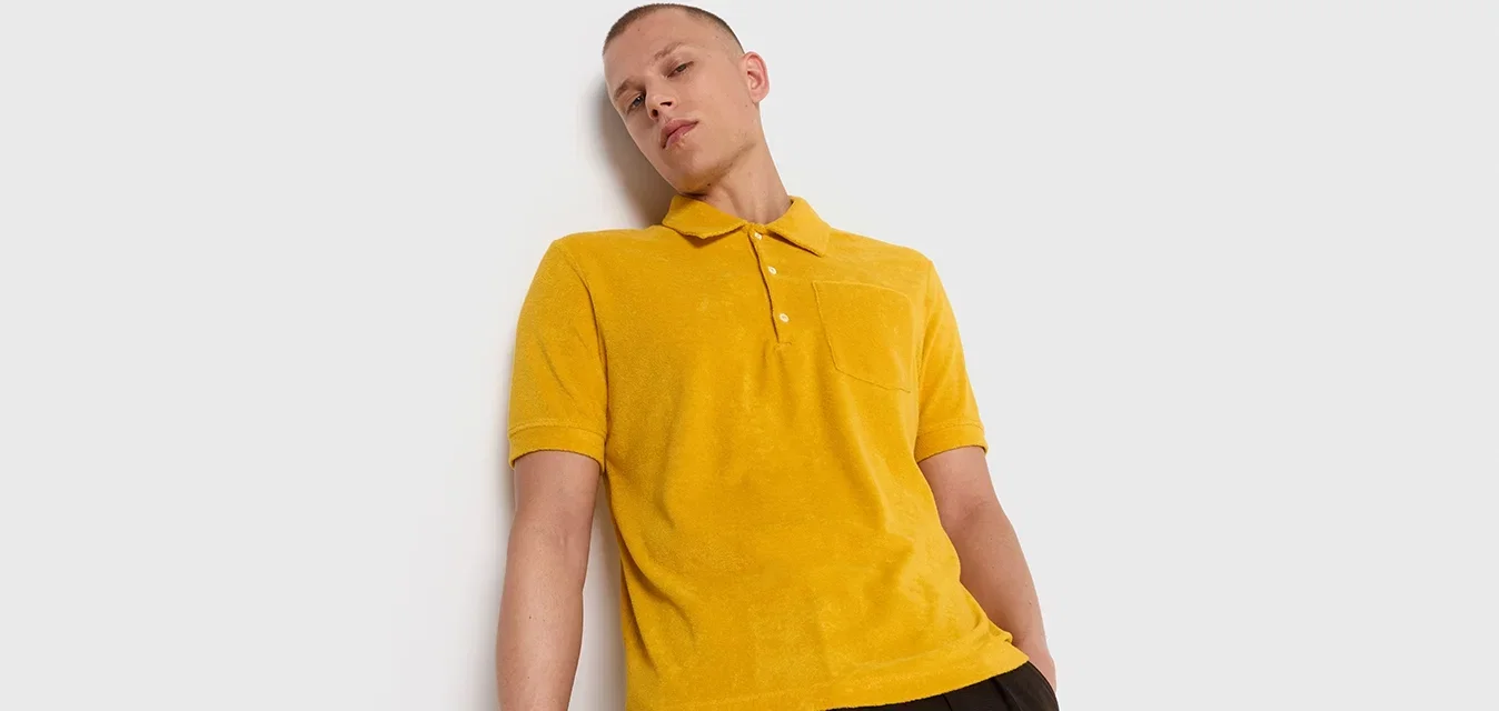 Geel polo shirt met een zachte textuur, korte mouwen en een drieknoopsluiting. Beschikt over een borstzak. Gepaard met losse donkere broeken.