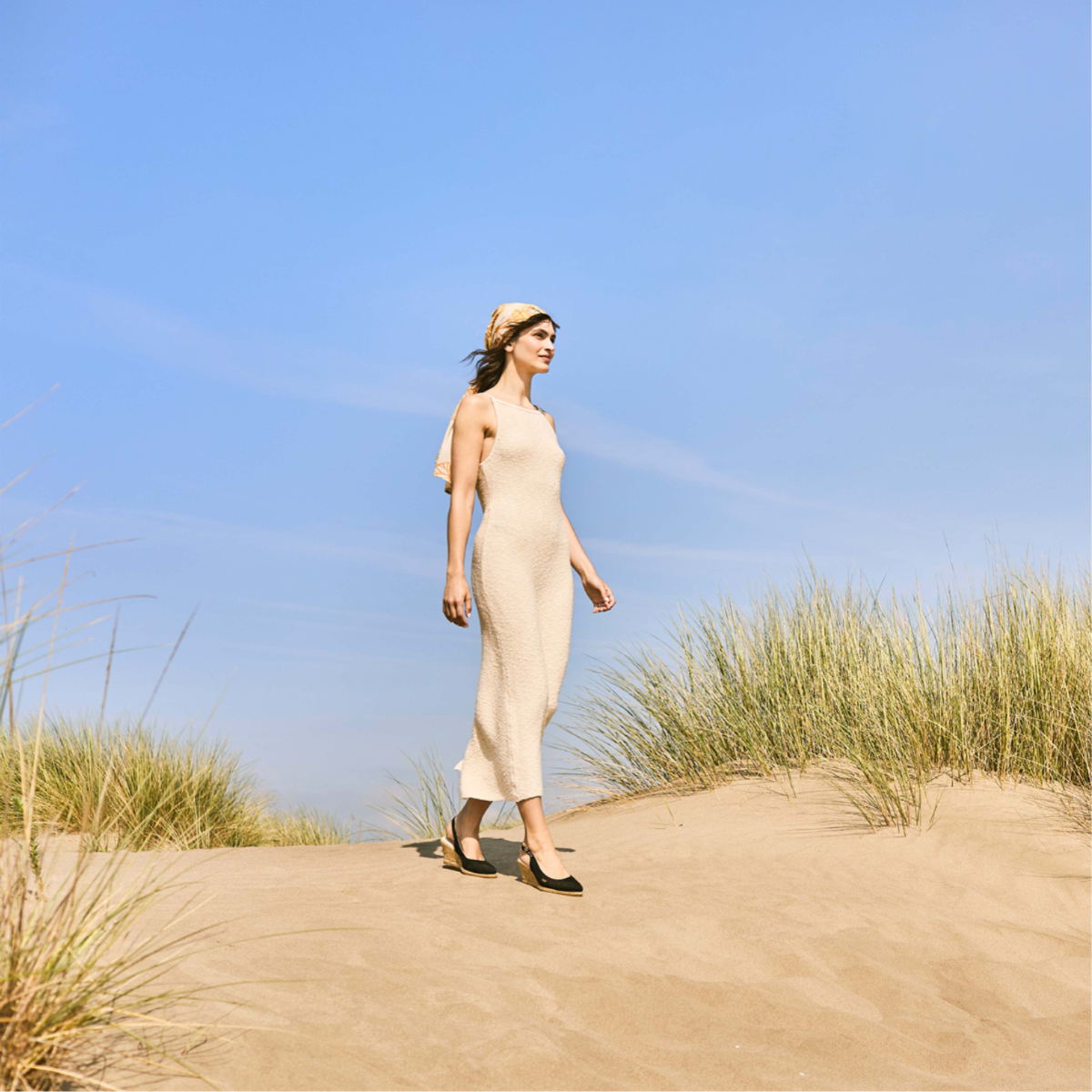 Woman walking across sandy dunes in a cream dress, headscarf, and black Viscata espadrilles, with “Travel Essentials” text over a clear blue summer sky.