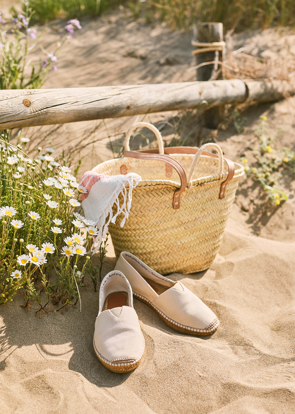 Beige Barceloneta espadrilles resting on warm sand beside a woven straw tote, beach cloth, and wild daisies in a soft summer beach setting.