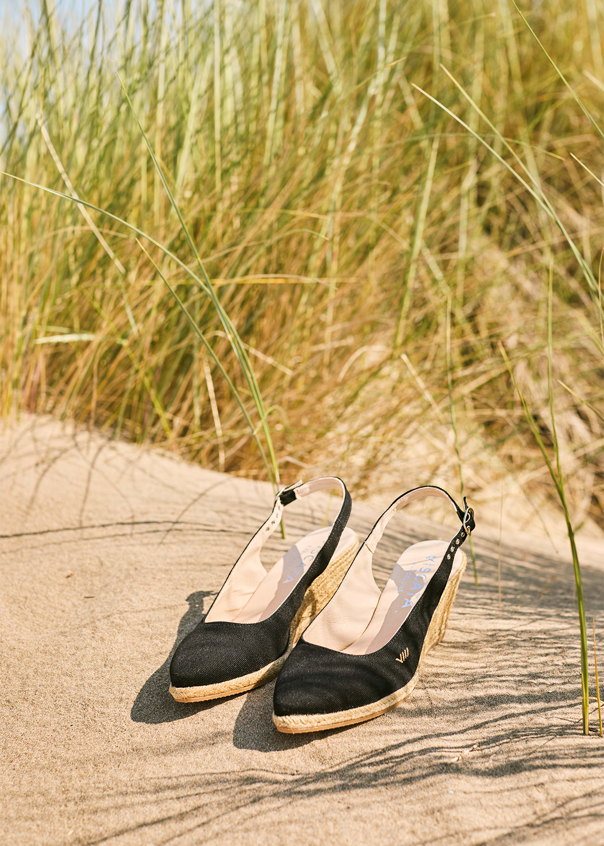 Black Palomera canvas slingback wedge espadrilles resting on warm sand among beach grass, with pointed toes, adjustable straps, and natural jute wedges.
