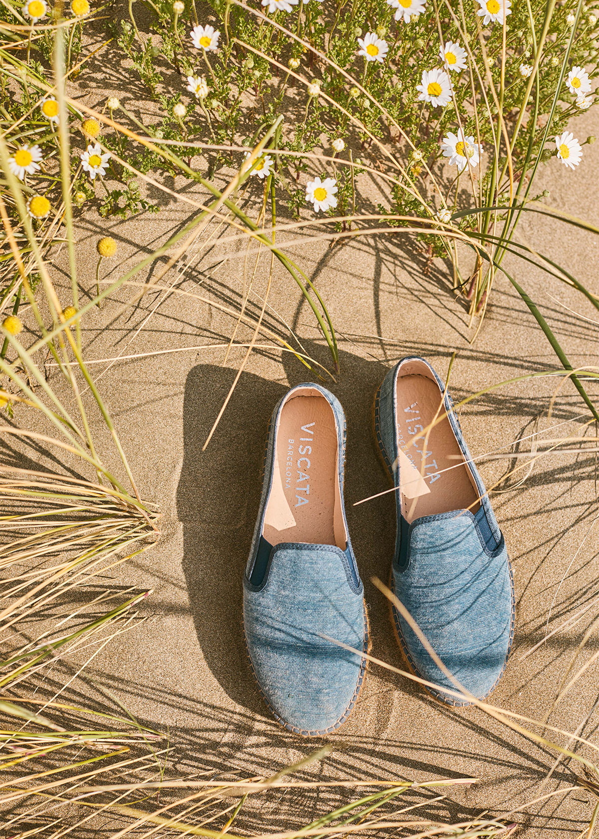 Washed denim Castell platform espadrilles resting on warm sand among beach grass and wild daisies, with natural jute soles and Viscata-branded insoles.