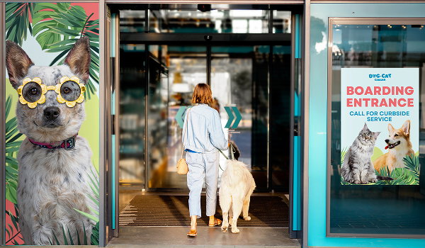 A woman walks a dog into a pet boarding and grooming business that has a window graphic and poster at the entryway.