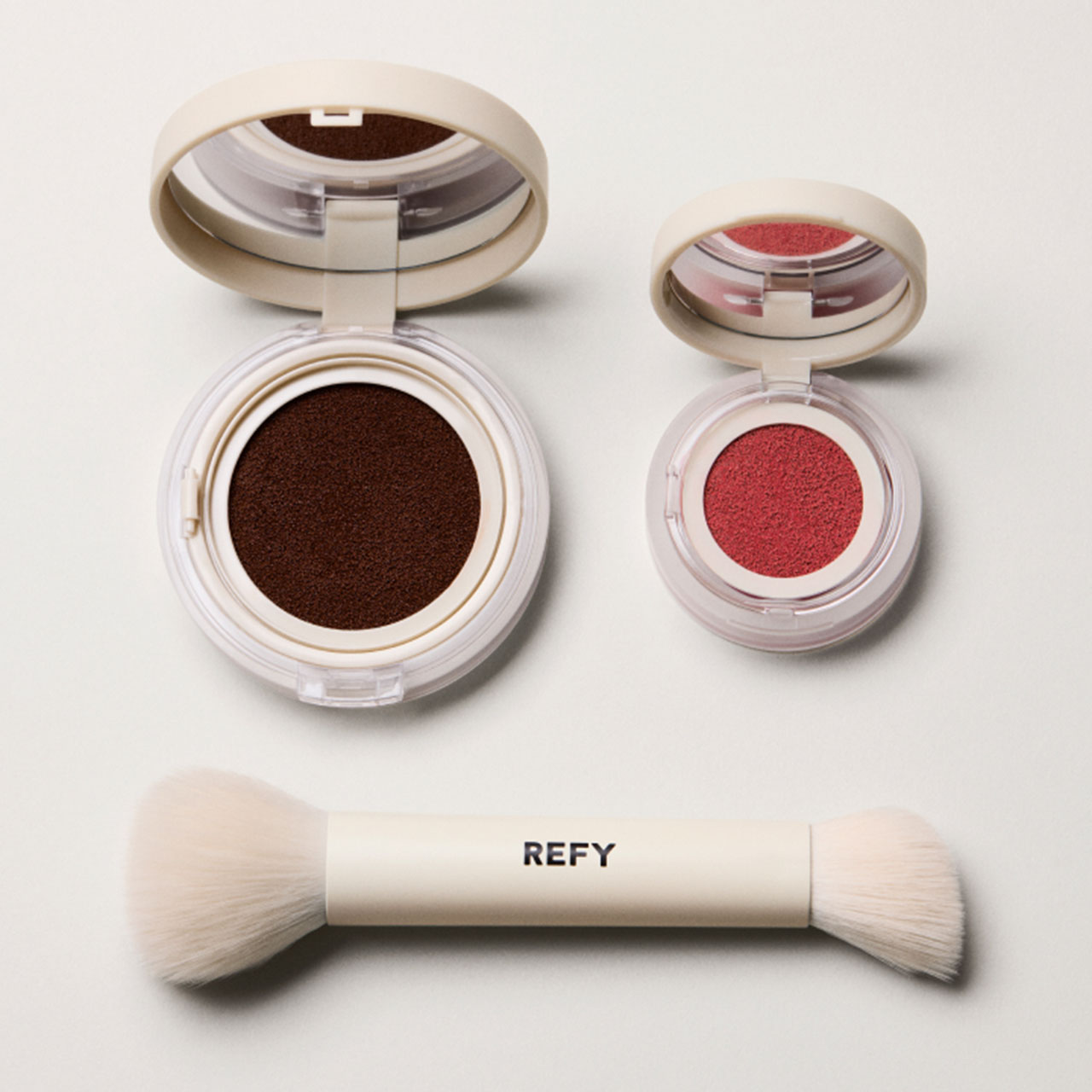 Refy makeup set with a makeup brush, a brown compact, and a pink eyeshadow in round containers on a neutral background.
