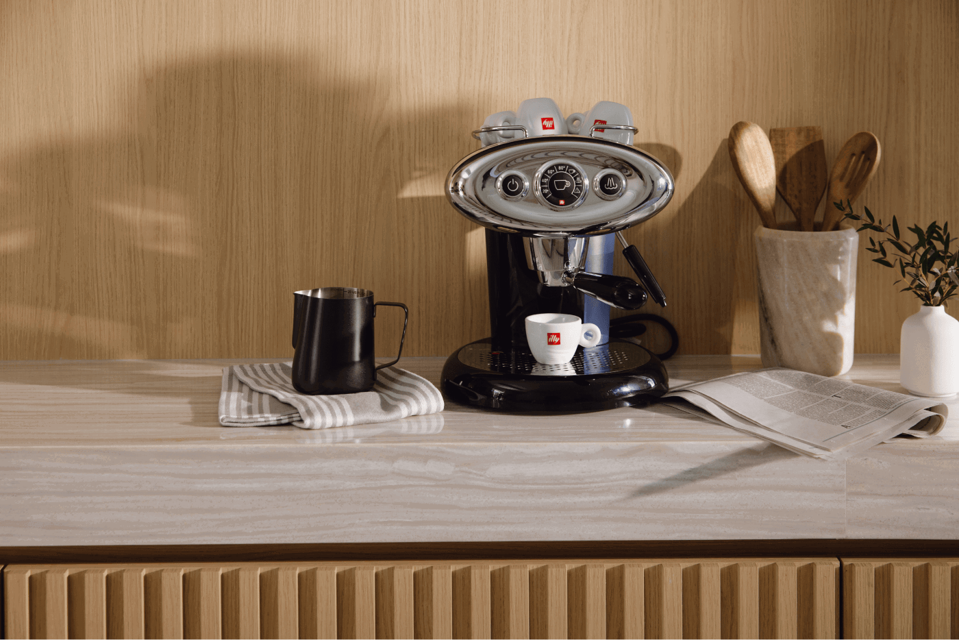 Espresso machine on a modern kitchen countertop with a coffee cup, milk pitcher, and wooden utensils