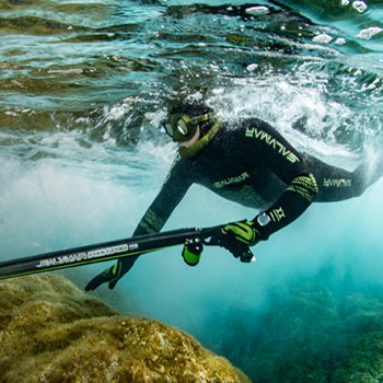 Freediver in black wetsuit using speargun near rocky reef under clear turquoise water
