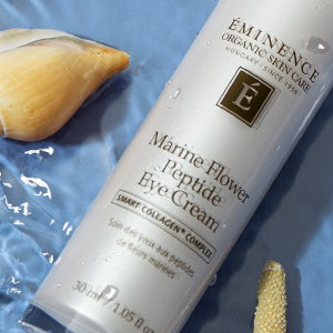 Eminence Organics bottle laying in rippling water next to a yellow seashell