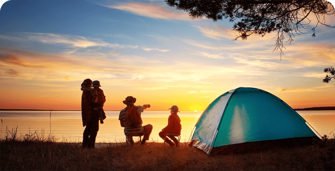 A family watches the sunset across a lake next to their tent.