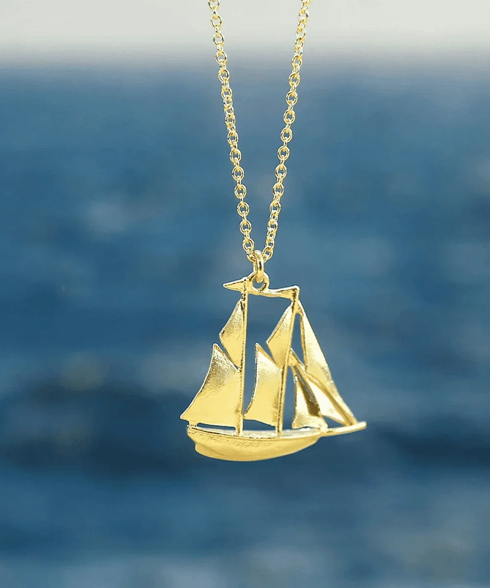 Alex Monroe Gold Galleon Ship Necklace in front of blue ocean