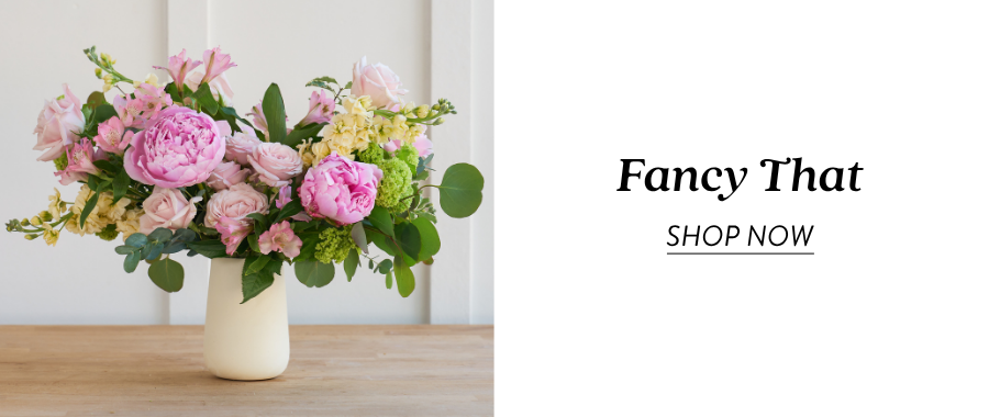 Product feature for “Fancy That,” showing a pink, peach, green, and white floral arrangement in a white vase on a wooden tabletop. Text on the right reads “Fancy That” with a “Shop Now” link.
