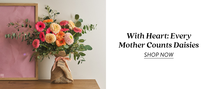 Product feature for “With Heart: Every Mother Counts Daisies,” showing a colorful burlap-wrapped bouquet with peach, yellow, pink, and orange flowers displayed on a wooden table beside a pink backdrop. Text on the right reads “With Heart: Every Mother Counts Daisies” with a “Shop Now” link.