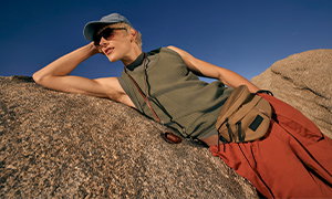 A man lies on some rocks under a blue sky. He wears a blue cap, a green sleeveless top, orange trousers, and a brown bumbag.