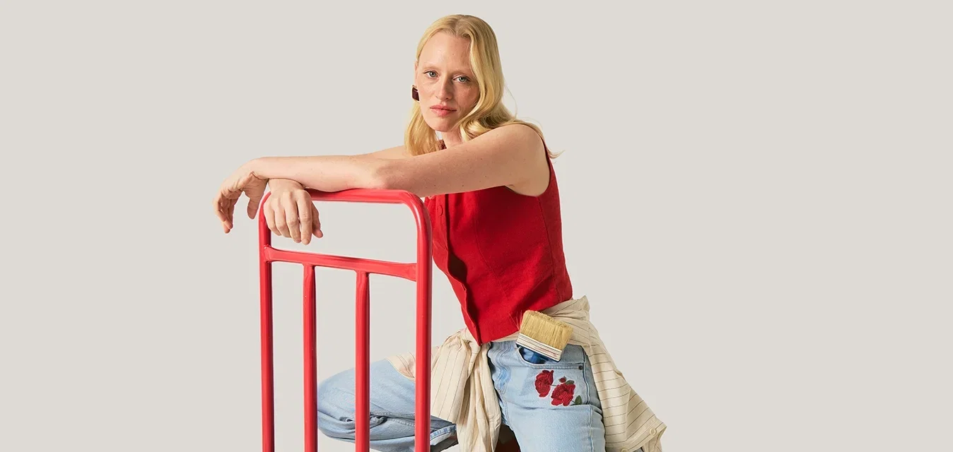Frau in rotem Oberteil und Jeans kniet auf einem schwarzen Plattformwagen mit rotem Griff vor neutralem Hintergrund.