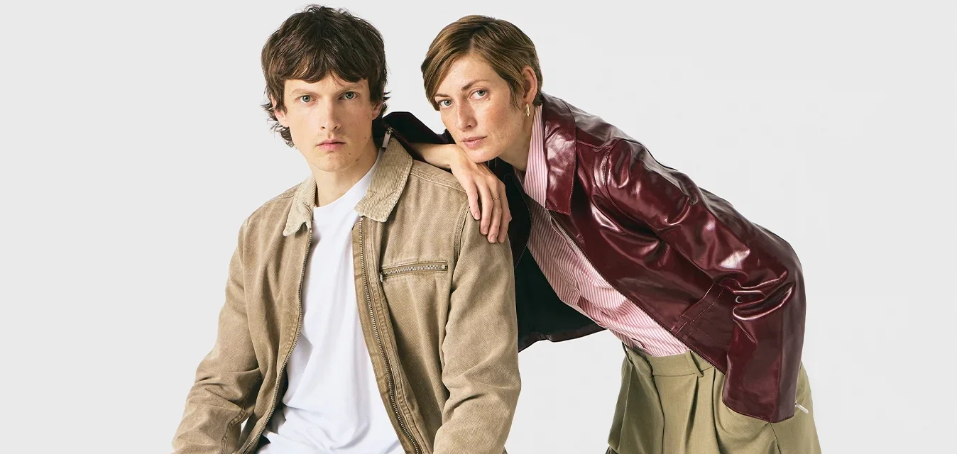 Man die op een kruk zit, gekleed in een beige jacket en zwarte broek; vrouw die op zijn schouder leunt, gekleed in een bordeauxkleurig jacket en kaki shorts.
