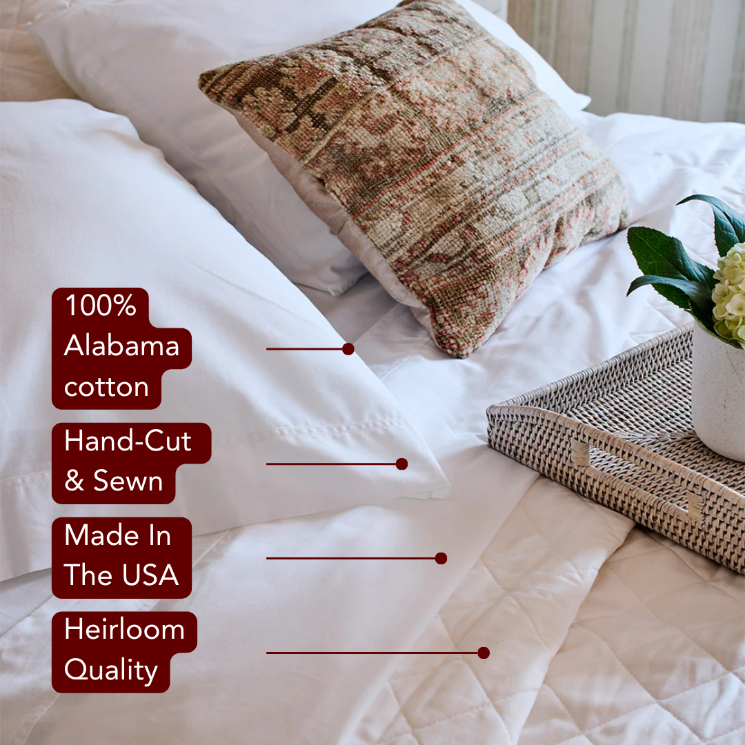 A close-up of a bed with crisp white sheets, a decorative pillow, and a wicker tray with a vase. Red overlay badges list product features like 100% Alabama cott