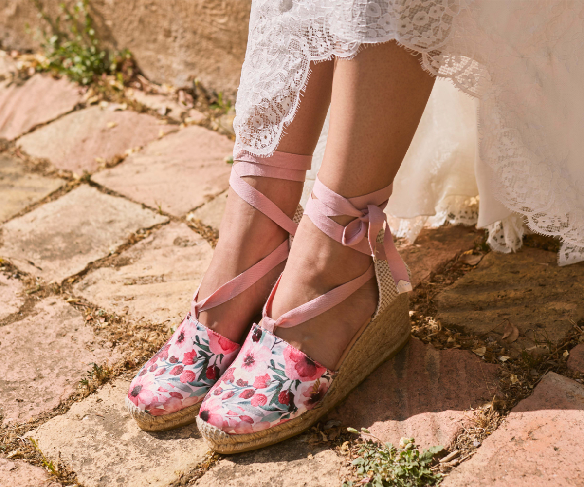 Close-up of floral Viscata espadrille wedge sandals with pink ankle ties, worn with a white lace dress on a sunlit stone path.