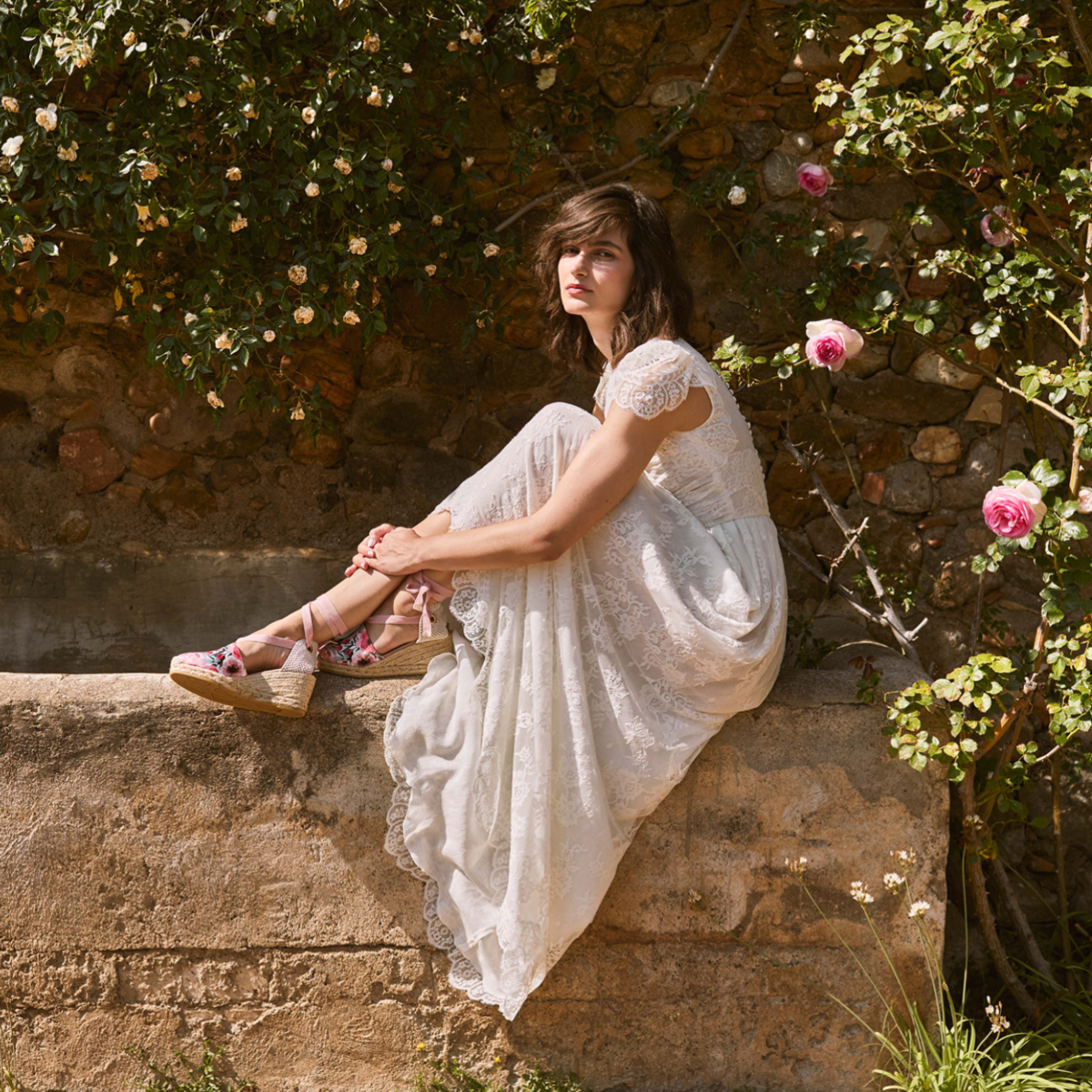 Woman in a white lace dress sitting on a sunlit stone wall surrounded by roses, wearing floral Viscata lace-up espadrille wedges.