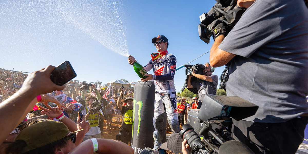 Jett Lawrence in 100% Armega goggles and Honda HRC gear celebrating a victory on the podium, spraying champagne to a cheering crowd at the 2026 Pro Motocross Championship