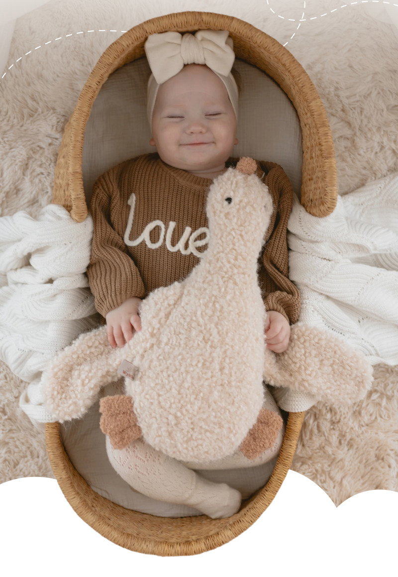 Smiling baby lying in a woven basket while holding a cream plush duck toy and wearing a personalized knit sweater.