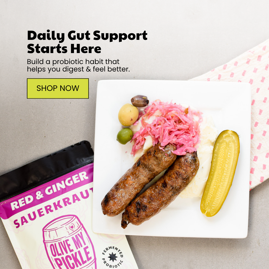 Overhead shot of a meal with grilled sausages, pink sauerkraut, olives, mashed potatoes, and a pickle, next to a 'Red & Ginger Sauerkraut' product package.