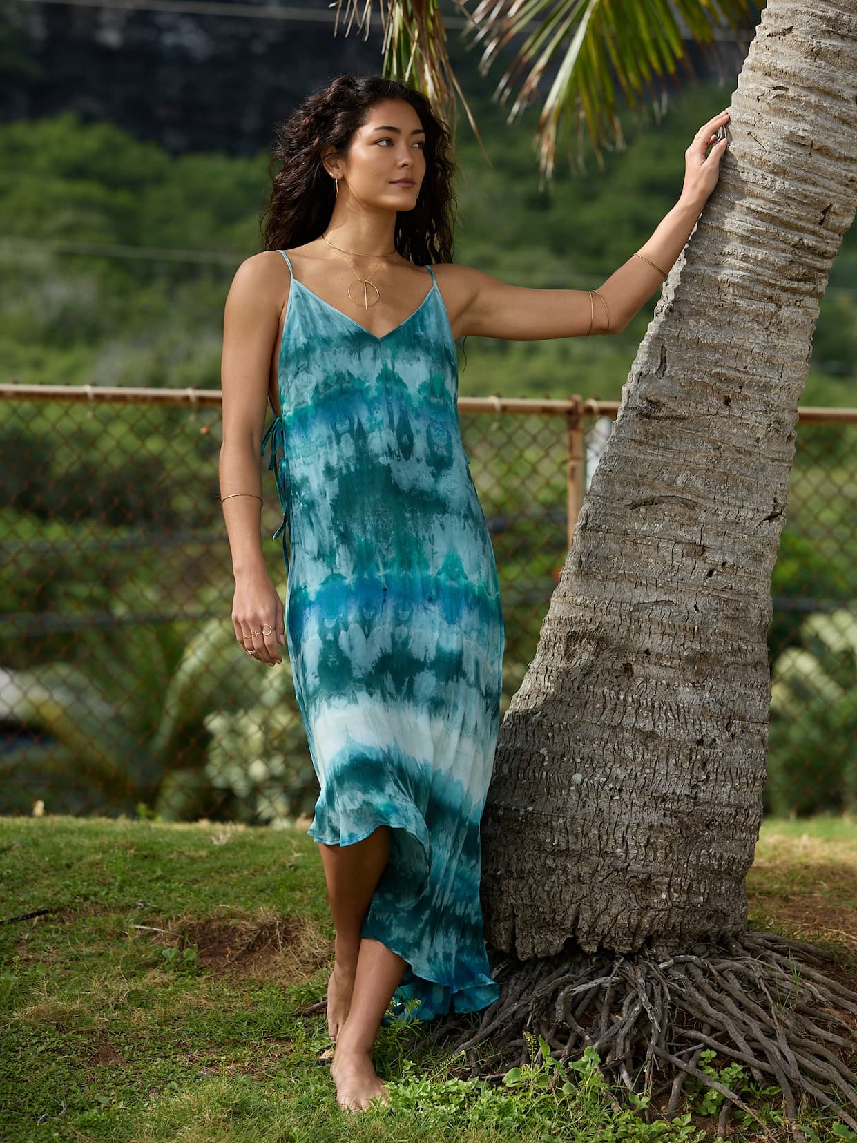 Woman wearing teal tie-dye dress leaning against a palm tree