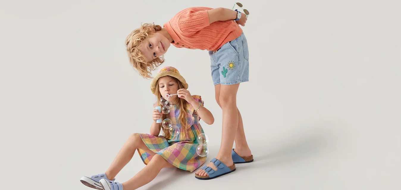 Twee kinderen, de een zittend zeepbellen blazend en de ander staand en voorover leunend nieuwsgierig naar de bellen kijkend.
