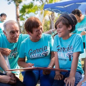 Multi-generational family smiles together at an outdoor gathering, wearing matching bright turquoise custom Garcia Family Reunion t-shirts.