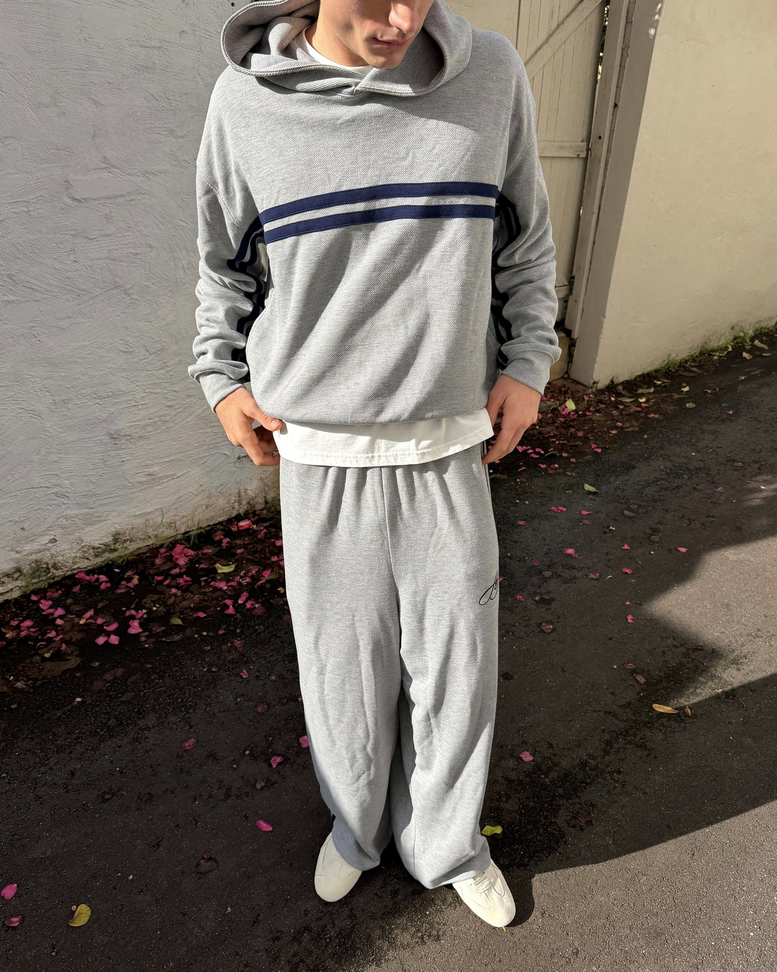 A medium shot captures a person standing outdoors wearing a grey hooded sweatshirt with navy blue stripes across the chest, a white t-shirt underneath...