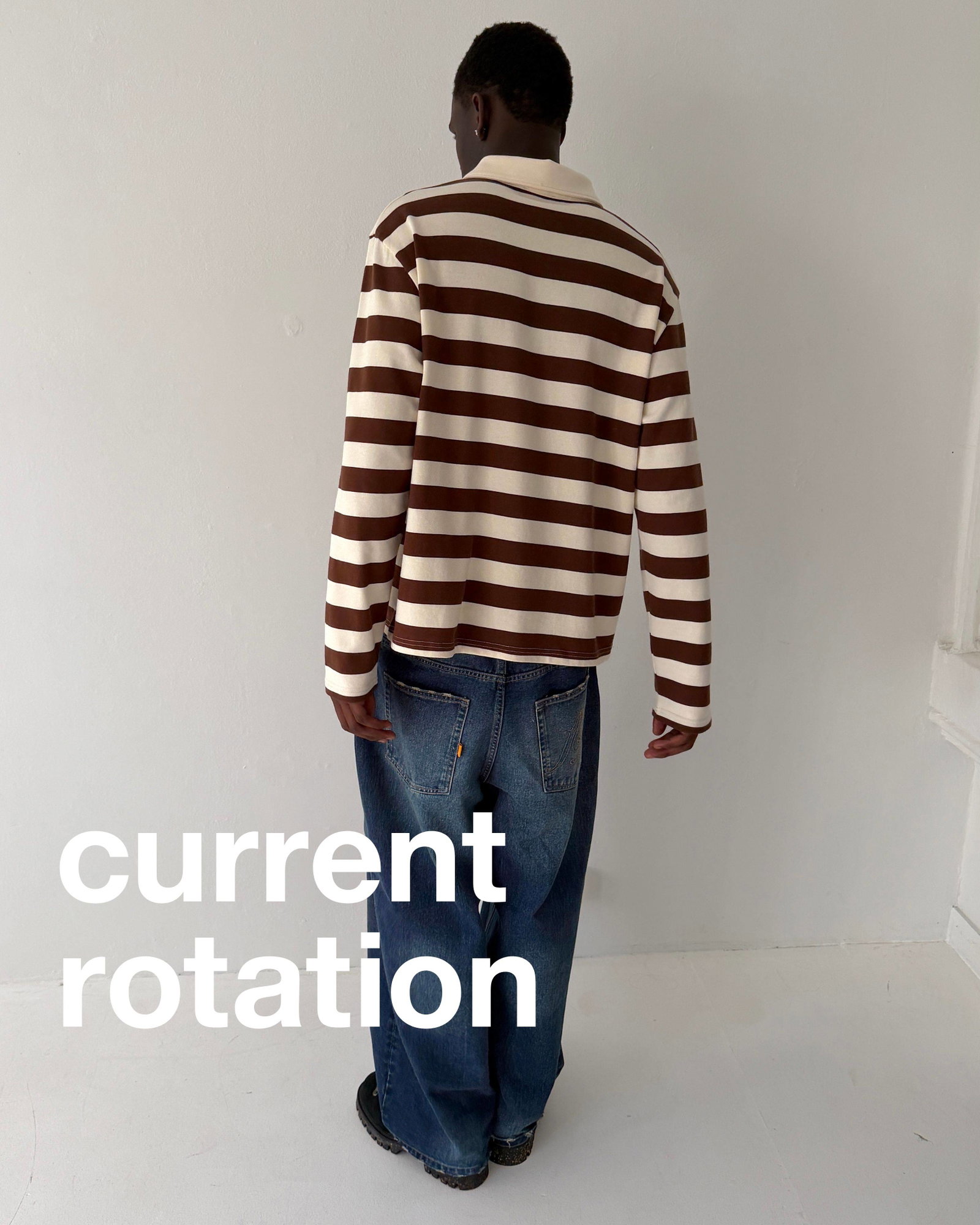 A medium shot shows a person from behind wearing a brown and cream horizontally striped long-sleeved collared shirt and loose-fitting blue jeans, with...