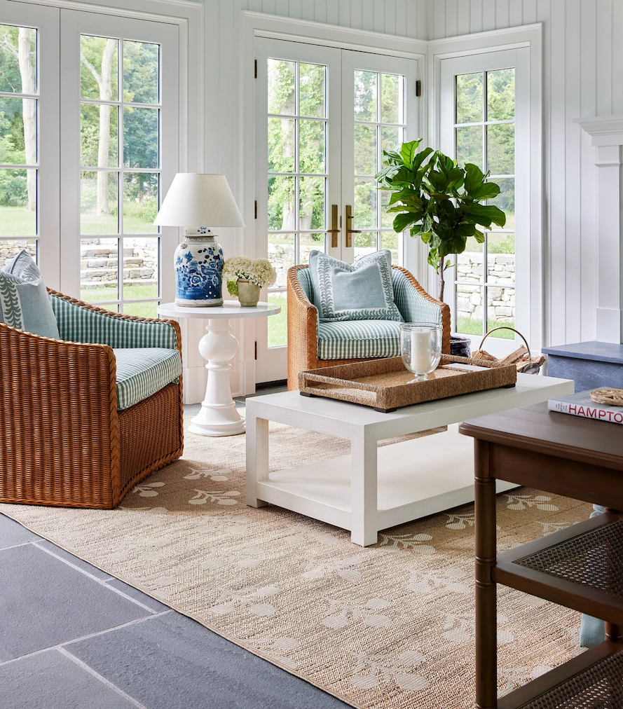 Bright living room with block pattern rug, white coffee table, woven side chairs, and side table with lamp and flowers on top.