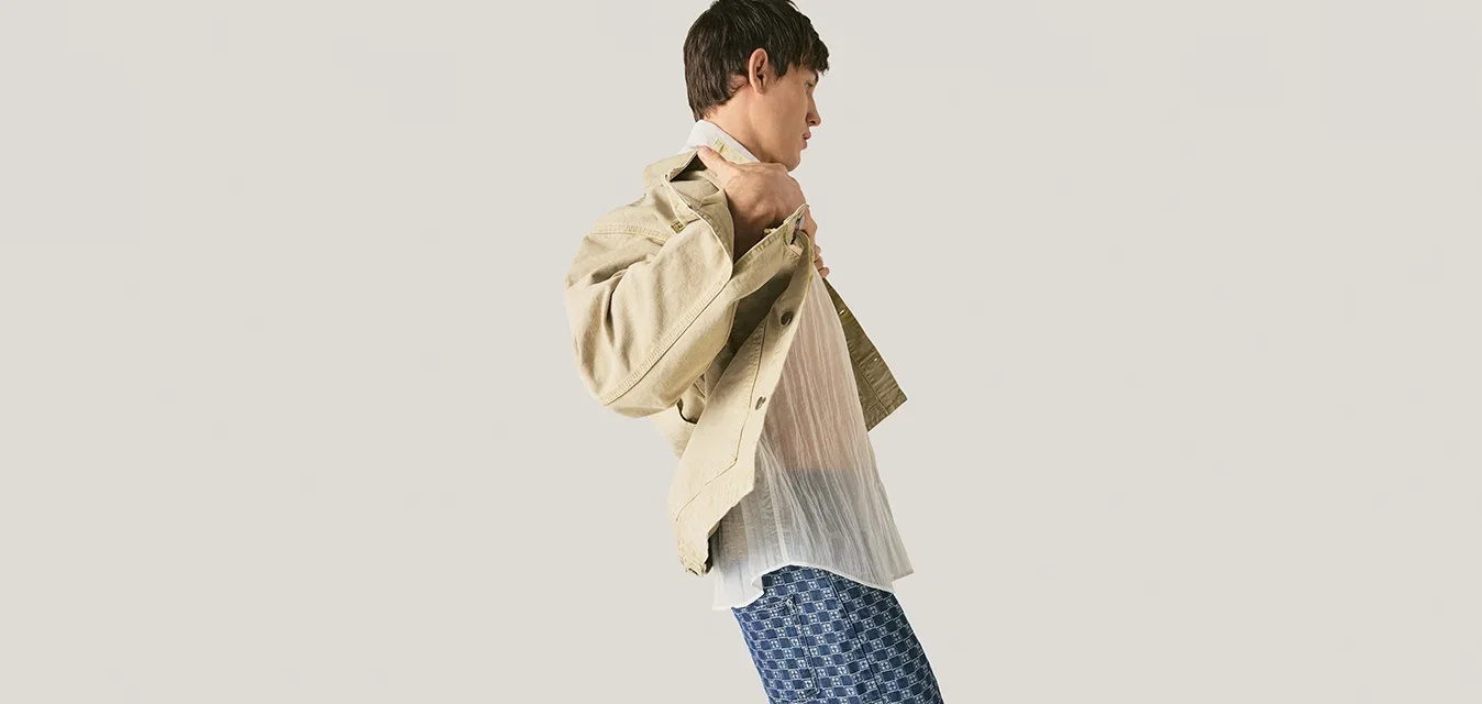 Junger Mann, der eine beige Jacke über einem weißen Hemd anpasst, trägt blaue gemusterte Hose und steht vor einem schlichten hellgrauen Hintergrund.