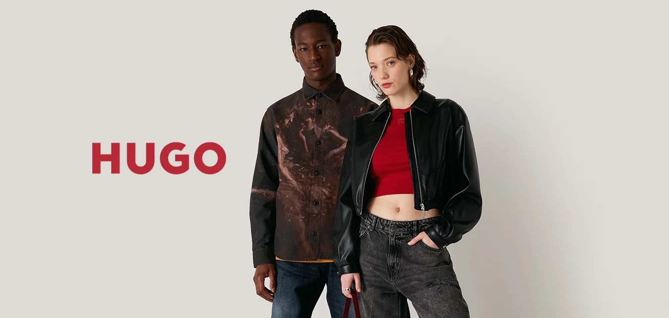 Zwei Models stehen vor einem neutralen Hintergrund. Eines trägt ein gemustertes braunes Hemd und blaue Jeans, das andere eine schwarze cropped Jacke und graue Hosen.
