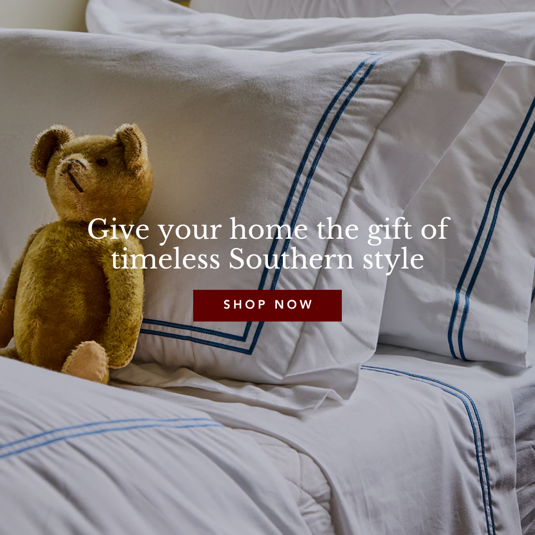 A teddy bear sitting on a neatly made bed with white sheets and pillows featuring blue embroidered lines.