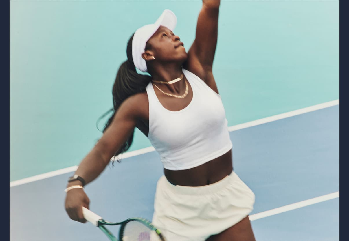 Vicky serving on a blue tennis court wearing Mejuri gold chain necklaces and a bracelet.