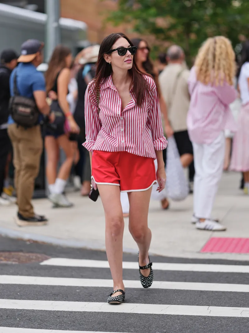 how to style running shorts - NEW YORK, NEW YORK - SEPTEMBER 12: A guest wears long dark brown straight hair parted in the center, black rectangular sunglasses, small gold hoop earrings, a shoulder bag with a gold chain strap and black body, a red-and-white vertically striped button-down shirt with rolled cuffs, red athletic shorts with white piped trim, and black studded Mary Jane flats with a single strap and flat soles, outside Area, during New York Fashion Week, on September 12, 2025 in New York, New York (Photo by Edward Berthelot/Getty Images)